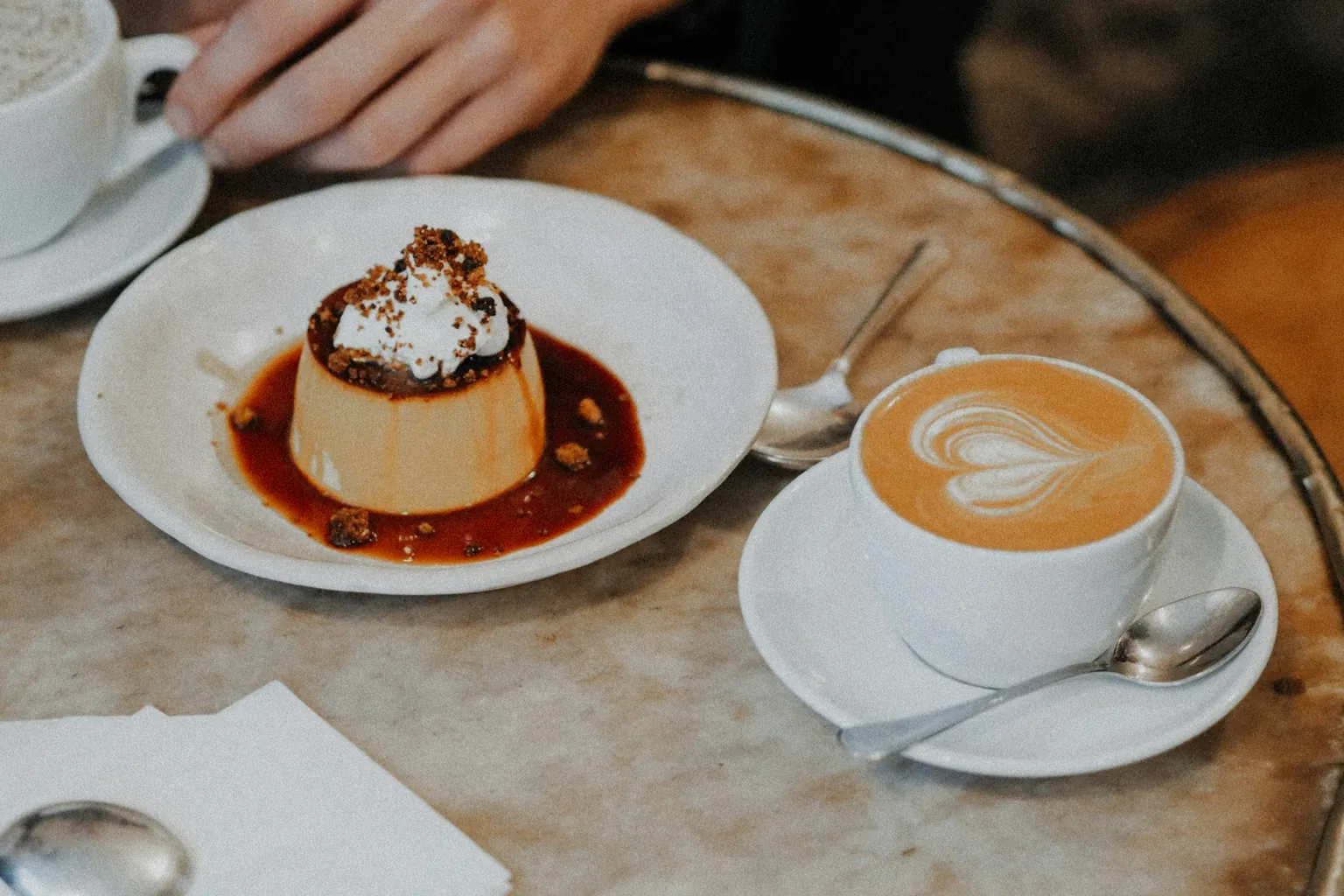 O cafea cu lapte cu inimă desenată în spumă și o cremă de zahăr ars, pe o masă rotundă de marmură dintr-o cafenea