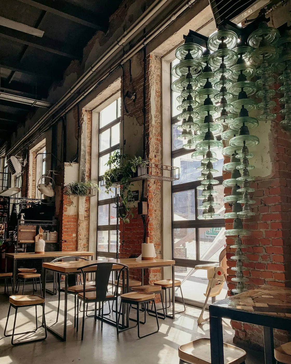 Sala unei cafenele cu pereți de cărămidă, ferestre mari, plante agățate și mese de lemn