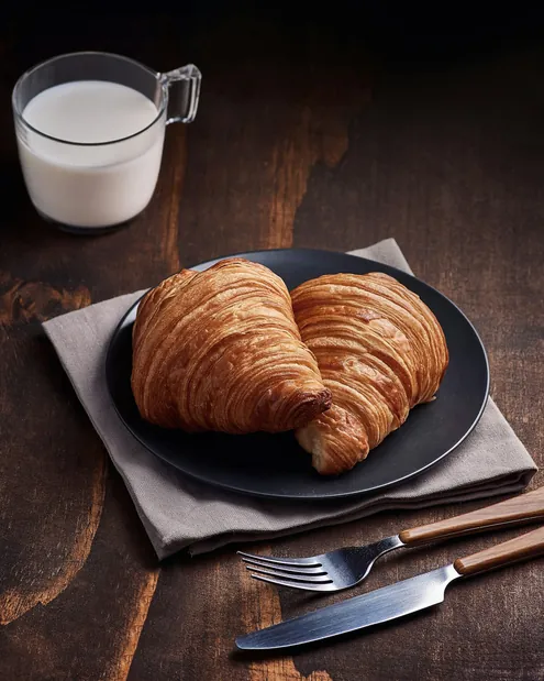 Două croissante pe o farfurie neagră, lângă un pahar cu lapte